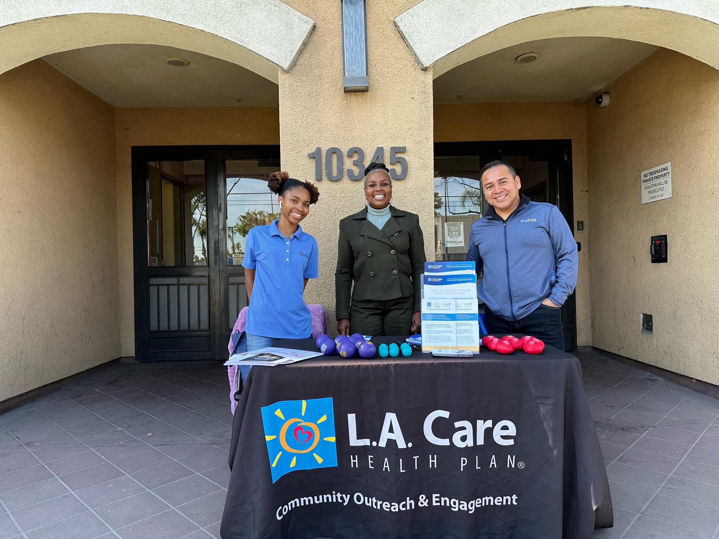 Two Health Matters Clinic team members and an L.A. Care representative standing behind an enrollment table set up outside the entrance of an apartment community