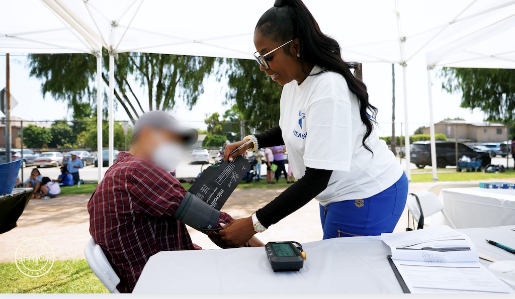 Health Matters Clinic team member fitting a blood pressure cuff on a seated community member at an outdoor pop-up table under a canopy